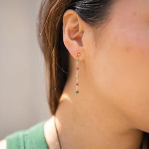 May include: A gold-tone drop earring featuring a delicate chain with five small, alternating round beads in shades of green and magenta. The earring is secured with a small gold-tone ball stud at the earlobe, creating a vertical, linear design.