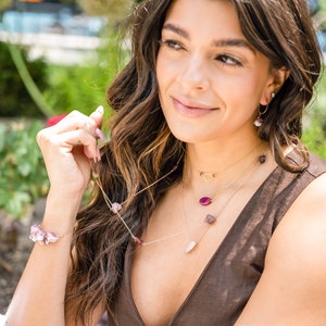 May include: A woman wearing a brown sleeveless top with a gold chain necklace featuring pink and red gemstone accents. She is also wearing a gold chain bracelet with pink gemstones and gold hoop earrings with a pink gemstone accent.