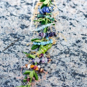 Peut inclure: Un ensemble de délicats colliers en or avec des accents de pierres précieuses variées, notamment des pierres bleues, vertes et violettes. Les colliers sont présentés sur une branche feuillue sur un fond gris texturé.