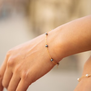 Puede incluir: Una delicada pulsera dorada adornada con pequeñas perlas de color gris oscuro. La pulsera se lleva en la muñeca, mostrando su diseño simple pero elegante. Las perlas están espaciadas uniformemente a lo largo de la cadena de oro, creando un brillo sutil.