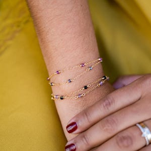 Peut inclure: Gros plan sur un poignet portant trois bracelets en chaîne dorée avec de petites perles violettes, bleues et noires.