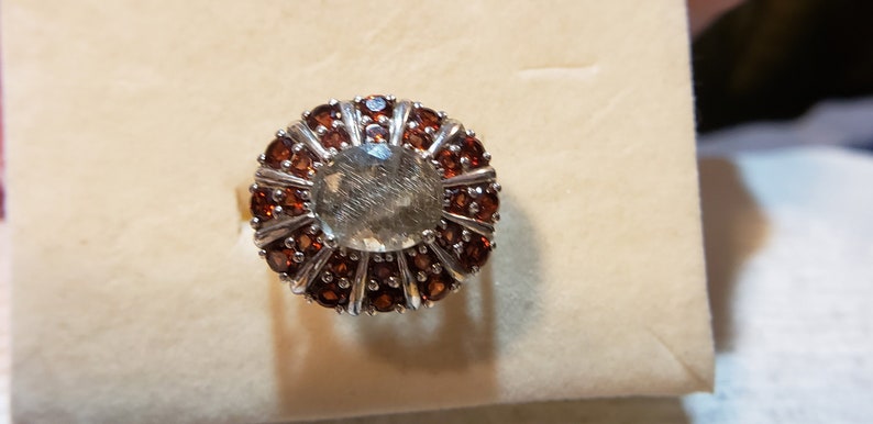 Centeral oval rutilated quartz surrounded by smaller and larger garnets in inner and outer stones Size 7. Platinum over sterling silver