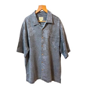 May include: A short-sleeved, button-down shirt in a muted blue-grey hue, adorned with a subtle floral pattern. The shirt features a chest pocket and is hanging on a wooden hanger. The fabric appears to be lightweight.