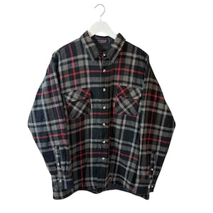 Puede incluir: Una camisa de franela a cuadros negra, gris y roja cuelga de una percha blanca. La camisa de manga larga tiene dos bolsillos en el pecho y una parte delantera abotonada. El patrón de cuadros presenta una mezcla de líneas grises oscuras, grises claras y rojas.