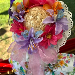 May include: A straw hat adorned with a vibrant arrangement of pink, orange, and purple artificial flowers, a small bird figurine, a white eyelet lace trim along the edge, and a sheer light purple ribbon tied into a bow at the front.