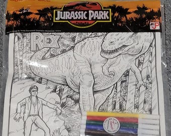 May include: A Jurassic Park doodle bag containing a black and white coloring sheet featuring a Tyrannosaurus Rex and a human figure in a jungle setting. The set includes five colored markers in yellow, red, green, blue, and brown.