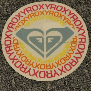 May include: A white round sticker with a blue and white logo of a heart with two arrows. The logo is surrounded by a circle of the word "ROXY" in pink, orange, and yellow.