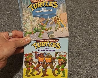 May include: Two Teenage Mutant Ninja Turtles books. The top book is titled The First Battle and shows a turtle in an alleyway. The bottom book is titled The Final Lesson and features all four turtles standing together with their signature weapons.