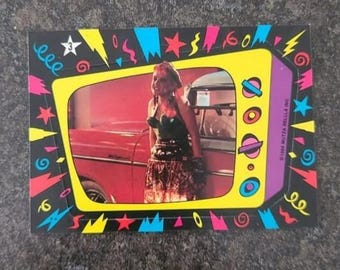 May include: A rectangular trading card featuring a yellow television-shaped frame with a purple side panel. Inside the screen, a person stands next to a red car. The border is black with colorful stars, squiggles, and the number 3 in a white star.