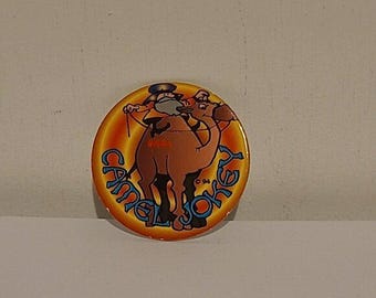 May include: A round, orange and yellow pin featuring a cartoon camel wearing a hat and holding a whip. The pin has the words "Camel Joke" in blue and orange lettering. The year "94" is printed on the pin.