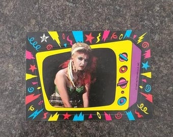 May include: A rectangular trading card featuring a photograph of a person with red hair and a headband inside a yellow television-shaped frame. The black border is decorated with colorful neon shapes including stars, lightning bolts, and swirls.