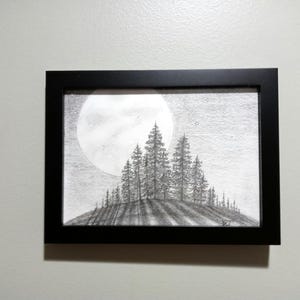 May include: A framed pencil drawing of a moonlit forest scene. The artwork features a large, bright moon in the background, with a silhouetted hill covered in tall pine trees in the foreground. The frame is black.