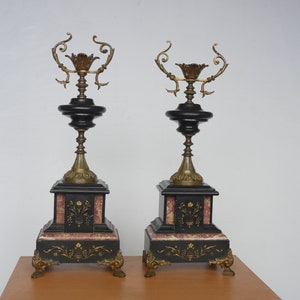 May include: A pair of black and gold candle holders with ornate details. The candle holders are made of marble and have a square base with a decorative design. The candle holders are on a wooden surface.