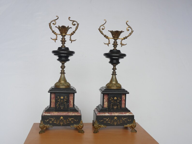 May include: A pair of black and gold candle holders with ornate details. The candle holders are made of marble and have a square base with a decorative design. The candle holders are on a wooden surface.