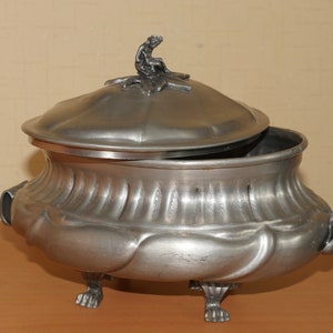 May include: A silver-toned metal tureen with a lid featuring a small seated figure finial. The bowl has a fluted upper section, a smooth rounded lower body, two side handles, and stands on three claw-style feet. The lid is slightly ajar.