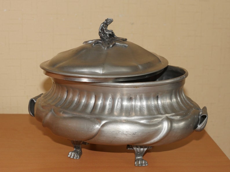 May include: A silver-toned metal tureen with a lid featuring a small seated figure finial. The bowl has a fluted upper section, a smooth rounded lower body, two side handles, and stands on three claw-style feet. The lid is slightly ajar.