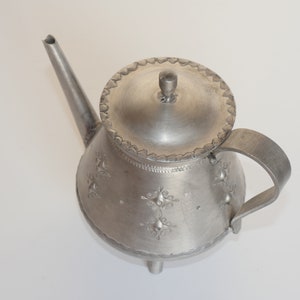 May include: A silver teapot with a lid and a handle. The teapot has a decorative design on the body and is sitting on a white surface.