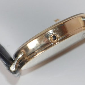 May include: A gold-tone wristwatch with a black leather band. The watch has a round face and a small, black dot on the side.