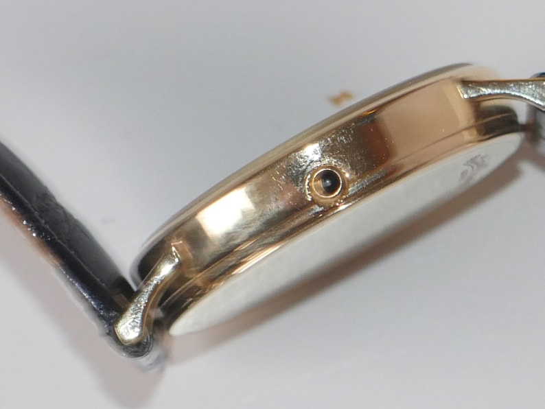 May include: A gold-tone wristwatch with a black leather band. The watch has a round face and a small, black dot on the side.