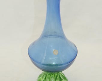 Könnte beinhalten: Eine blaue Glasvase mit bauchigem Körper und schlankem Hals. Die Vase steht auf einem grünen, strukturierten Sockel. Das Glas scheint transparent zu sein. Ein kleiner, runder Aufkleber ist auf dem Vasenkörper sichtbar.