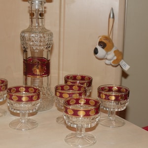 May include: Set of six clear glass stemmed shot glasses with a red and gold band around the top. The glasses are arranged on a white surface next to a clear glass decanter with a red and gold band around the middle.