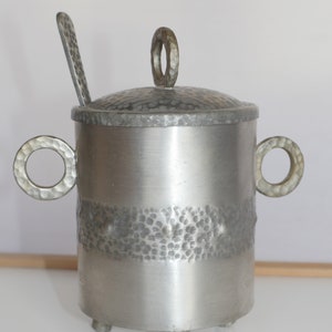 May include: Silver sugar bowl with a hammered finish and a lid with a ring handle. The bowl has two ring handles and sits on three feet.