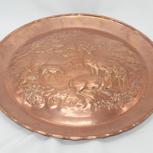 Puede incluir: Una bandeja decorativa redonda de color cobre con una escena en relieve detallada de ciervos en un entorno forestal. La bandeja tiene un borde festoneado y un acabado pulido, adecuada para exhibir.