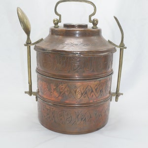 K&ouml;nnte beinhalten: Ein dreist&ouml;ckiger Tiffin-Beh&auml;lter aus Kupfer mit einem verzierten, gravierten Design. Der Beh&auml;lter hat einen Messinggriff und zwei Messingl&ouml;ffel, die an den Seiten befestigt sind.