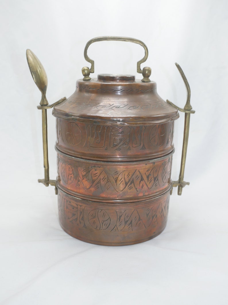 K&ouml;nnte beinhalten: Ein dreist&ouml;ckiger Tiffin-Beh&auml;lter aus Kupfer mit einem verzierten, gravierten Design. Der Beh&auml;lter hat einen Messinggriff und zwei Messingl&ouml;ffel, die an den Seiten befestigt sind.