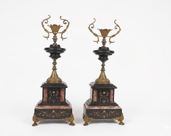 May include: A pair of tall decorative stands featuring black and reddish-marbled stone bases with gold-toned metal accents. Each stand has a square base with ornate feet, a central column, and a top section with curved metal scrollwork details.