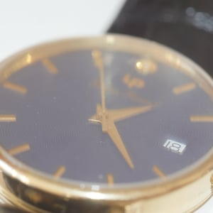 May include: A gold-tone wristwatch with a black face and gold-tone hands. The watch face has a date window at the 3 o'clock position and the number 18 is displayed.
