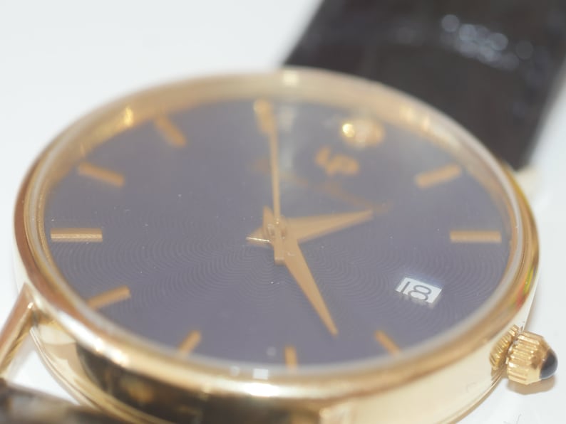 May include: A gold-tone wristwatch with a black face and gold-tone hands. The watch face has a date window at the 3 o'clock position and the number 18 is displayed.