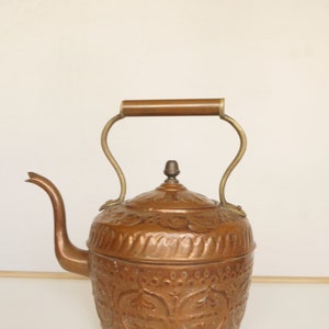Può includere: Un bollitore di rame con un design in rilievo decorato e un manico in legno. Il bollitore ha un beccuccio e un coperchio con un pomo.