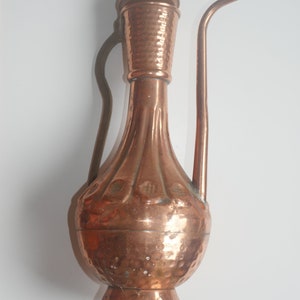 Op de afbeelding: Een gehamerde koperen kan met een tuit en een deksel. De kan heeft een decoratief ontwerp rond het midden en een brede basis.