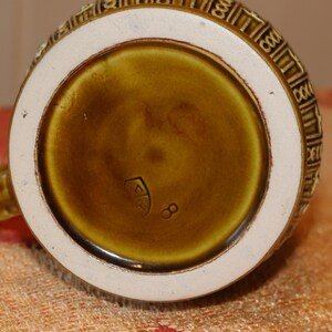 Może przedstawiać: Zbliżenie na sp&oacute;d brązowego ceramicznego kubka z białym rantem. Kubek ma ozdobny wz&oacute;r wok&oacute;ł rantu i znak producenta, kt&oacute;ry brzmi "ER 8".