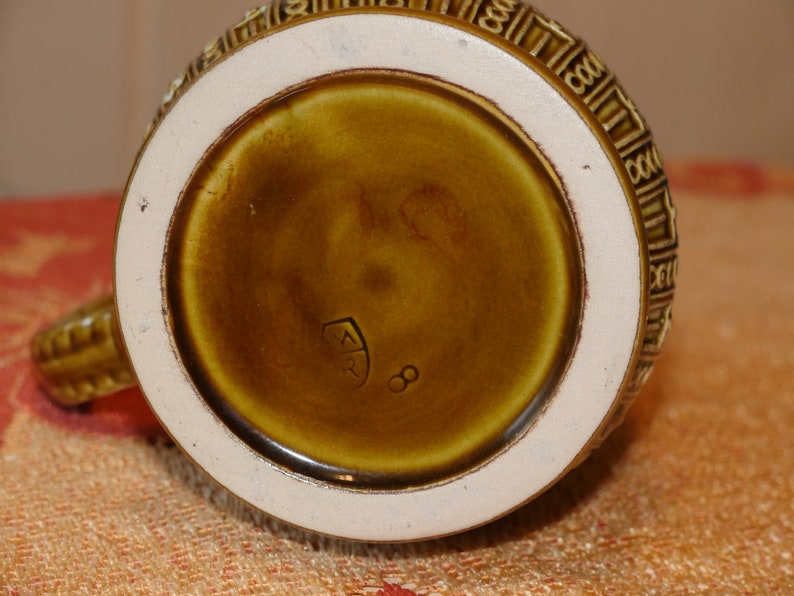 Może przedstawiać: Zbliżenie na sp&oacute;d brązowego ceramicznego kubka z białym rantem. Kubek ma ozdobny wz&oacute;r wok&oacute;ł rantu i znak producenta, kt&oacute;ry brzmi "ER 8".