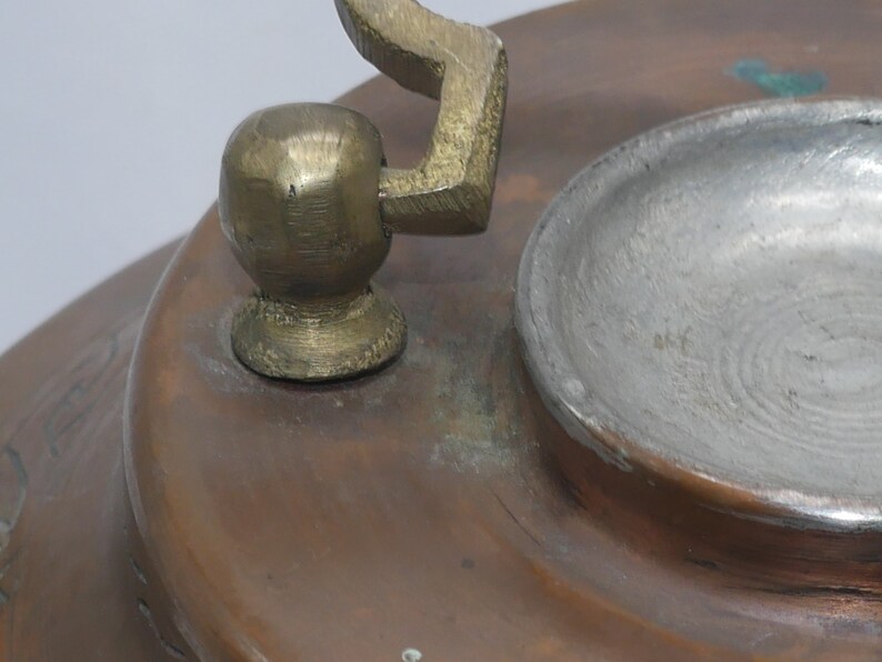 K&ouml;nnte beinhalten: Nahaufnahme eines Kupferkessels mit einem Messinggriff und -deckel. Der Deckel hat einen kleinen, runden Knopf oben und einen gebogenen Griff.
