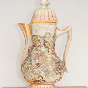 May include: A ceramic teapot with a detailed floral design and a pink and green lid. The teapot features a raised relief of people and trees.