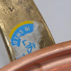 May include: Close-up of a copper pot with a brass handle. The handle has a blue and yellow sticker that reads "MADE IN SWEDEN".