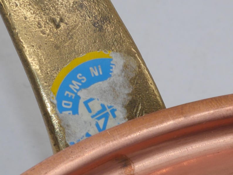 May include: Close-up of a copper pot with a brass handle. The handle has a blue and yellow sticker that reads "MADE IN SWEDEN".