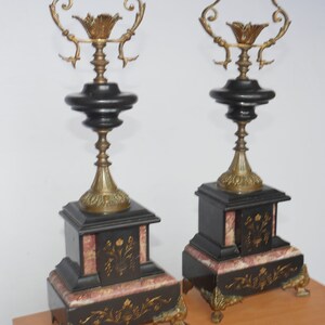 May include: A pair of black and gold decorative fireplace candle holders with ornate details. The bases are made of black marble with pink accents and gold leaf designs.