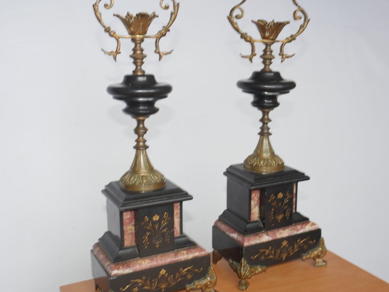 May include: A pair of black and gold decorative fireplace candle holders with ornate details. The bases are made of black marble with pink accents and gold leaf designs.