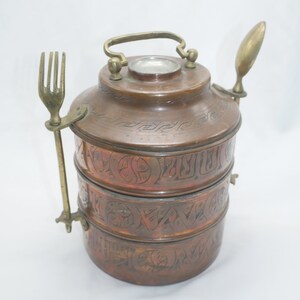 K&ouml;nnte beinhalten: Ein dreist&ouml;ckiger Tiffin-Beh&auml;lter aus Kupfer mit einem gravierten Design. Der Beh&auml;lter hat einen Messinggriff und wird mit einer passenden Gabel und einem L&ouml;ffel geliefert.