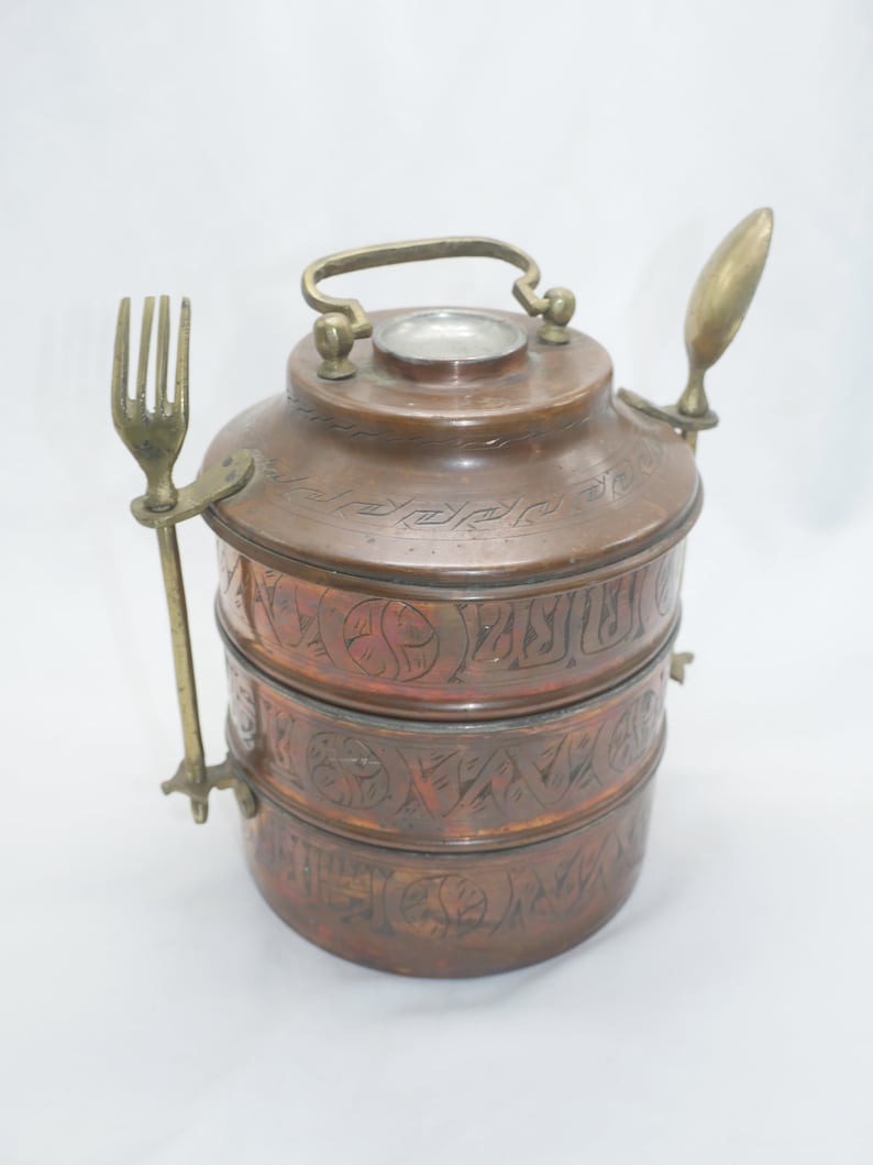 K&ouml;nnte beinhalten: Ein dreist&ouml;ckiger Tiffin-Beh&auml;lter aus Kupfer mit einem gravierten Design. Der Beh&auml;lter hat einen Messinggriff und wird mit einer passenden Gabel und einem L&ouml;ffel geliefert.