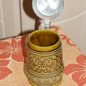 Może przedstawiać: Brązowy ceramiczny kufel do piwa z pokrywką. Kufel zdobią misterne złote i brązowe wzory oraz srebrna pokrywka.