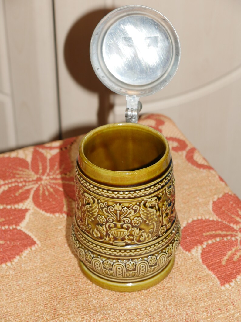 Może przedstawiać: Brązowy ceramiczny kufel do piwa z pokrywką. Kufel zdobią misterne złote i brązowe wzory oraz srebrna pokrywka.