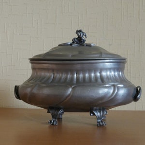 May include: A dark gray metal tureen with a rounded body, featuring a fluted design and two side handles. The lid is topped with a small sculptural finial, and the base is supported by four ornate feet that extend from the bottom of the vessel.