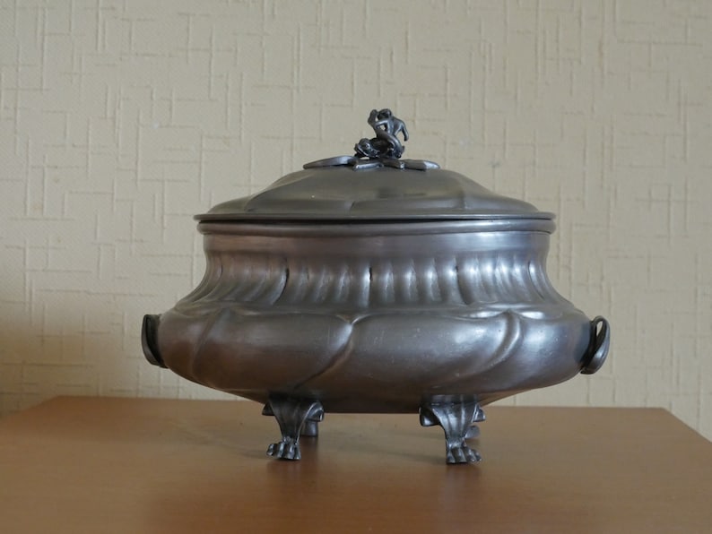 May include: A dark gray metal tureen with a rounded body, featuring a fluted design and two side handles. The lid is topped with a small sculptural finial, and the base is supported by four ornate feet that extend from the bottom of the vessel.