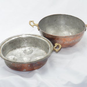 Op de afbeelding: Twee koperen potten met messing handvatten. De kleinere pot heeft drie inkepingen op de bodem.