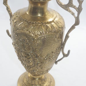 May include: A gold metal vase with intricate floral designs and two ornate handles. The vase has a round base and a tall, slender body.